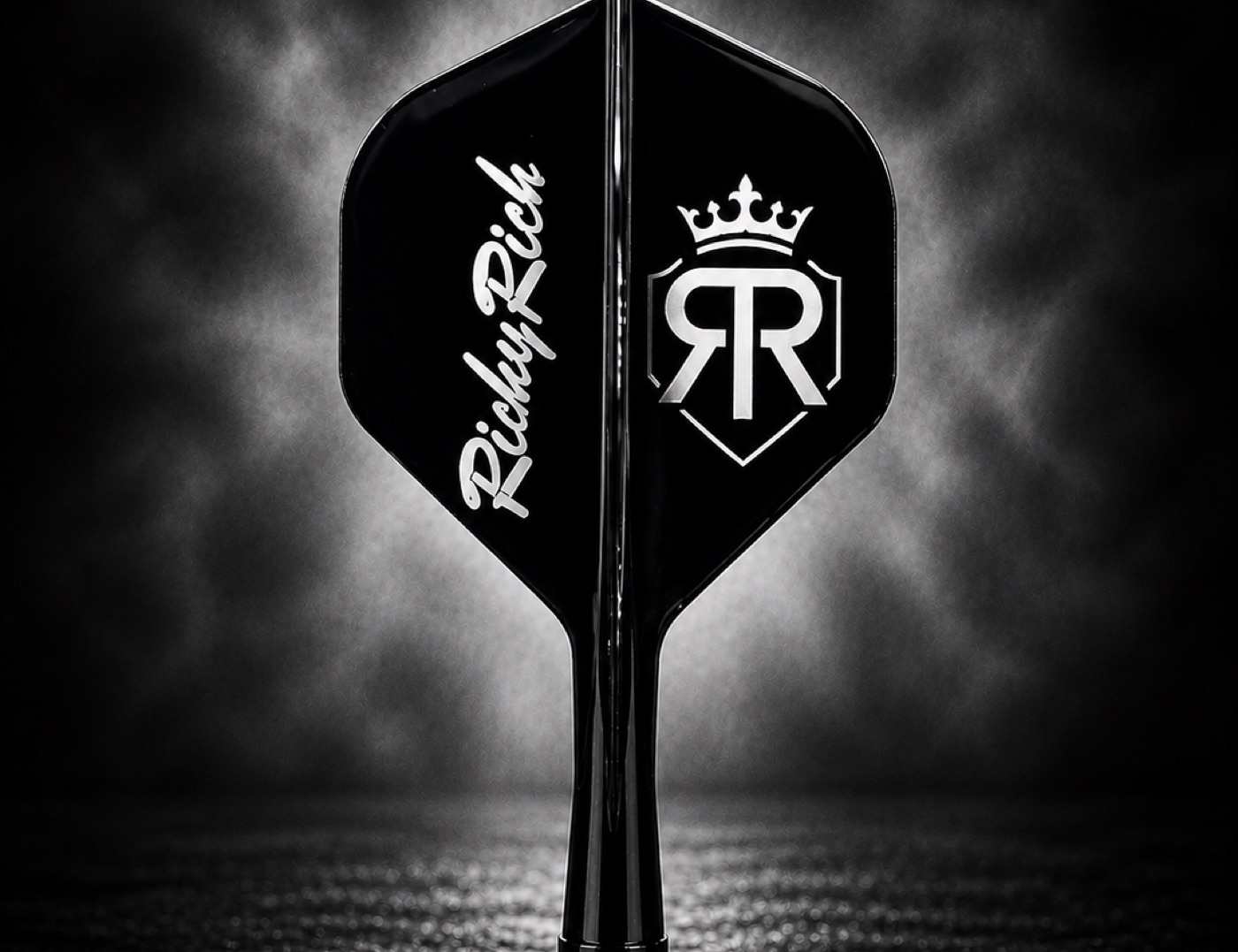 A black dart flight engraved in white with a name in script and a crowned monogram crest, lit against a dark smoky background