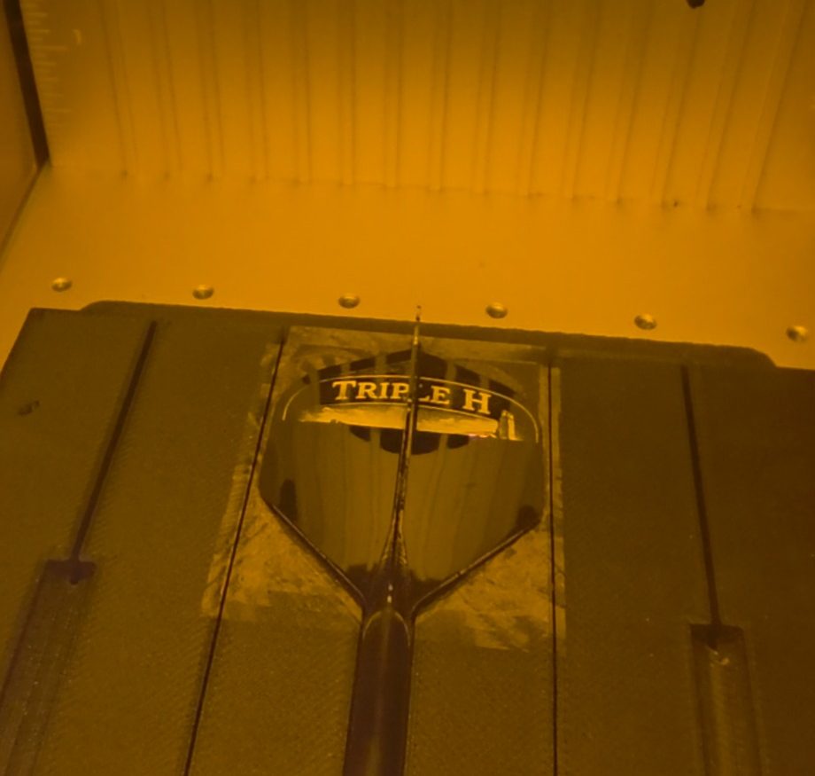 A dart flight held in the bed of a laser engraving machine, lit orange, with a name being cut into its surface